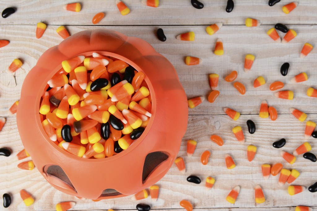 High angle shot of a plastic halloween pumpkin filled with candy on a white rustic wood table. Horizontal format with candy corn and jelly beans scattered on the surface.