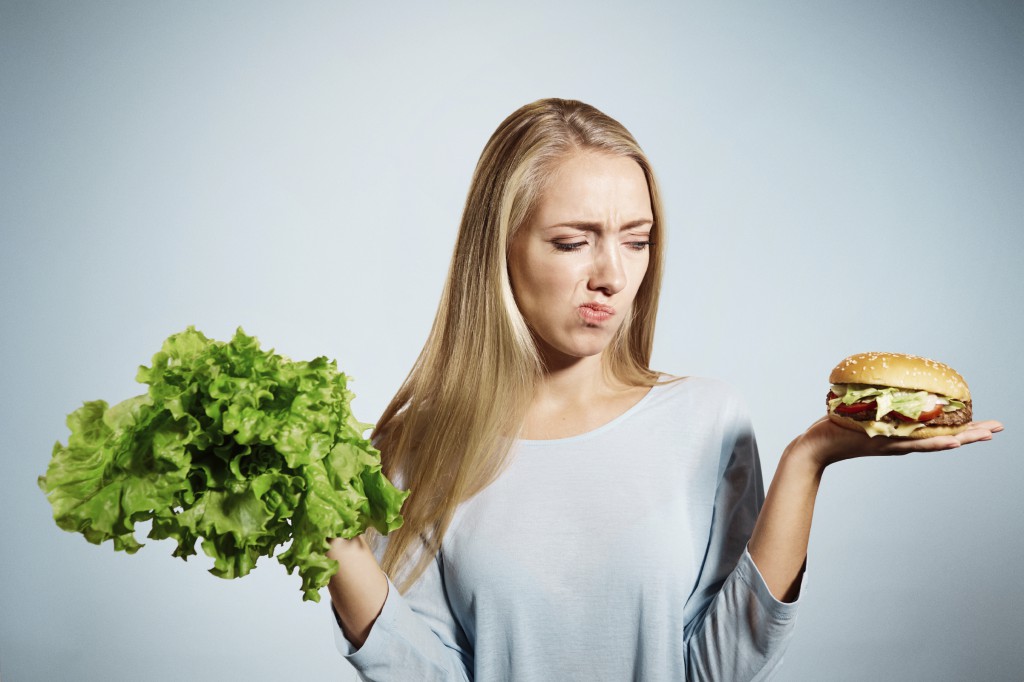 Pensive woman making decision between healthy food and fast food, over blue background