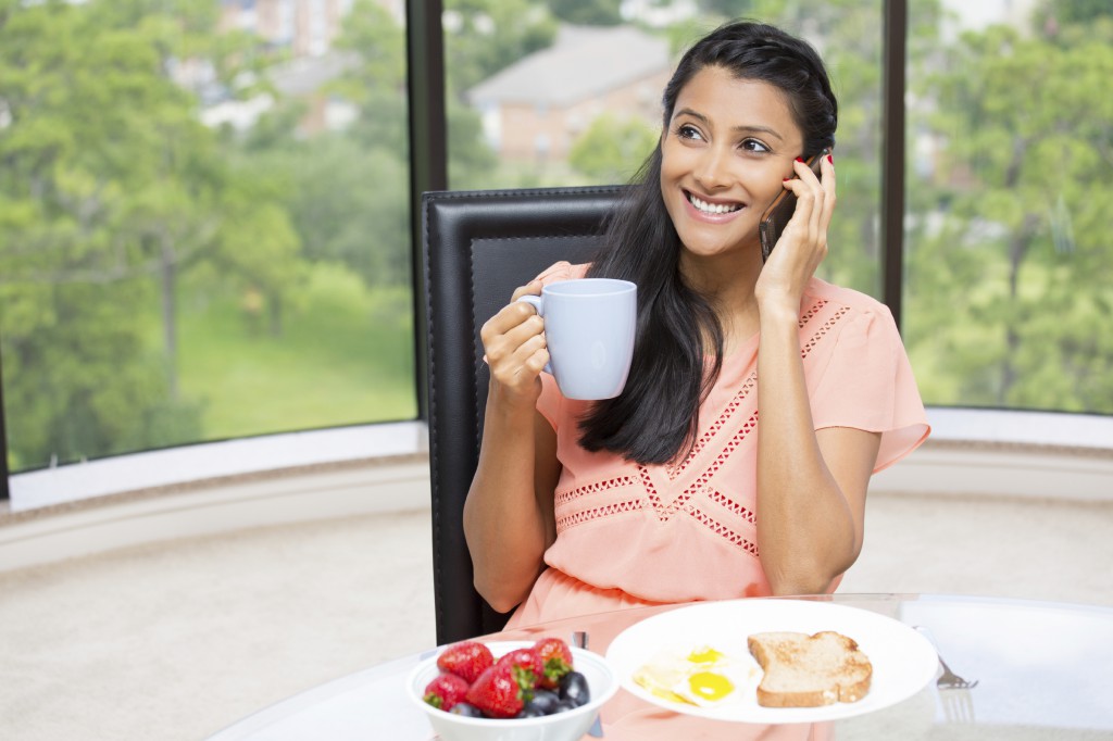 Closeup portrait of a young, attractive businesswoman, kick start day, health breakfast, fruit bowl, egg ,green tea, smiling, talking over the phone. Isolated glass window indoor greenery background.