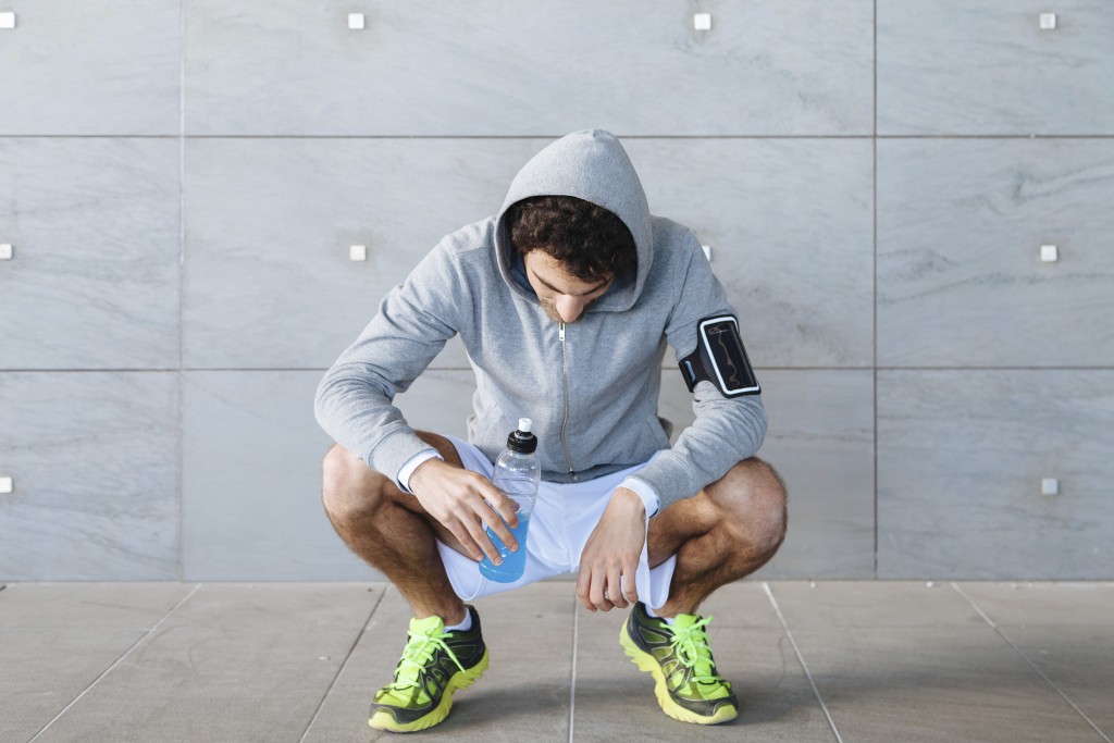 A young man in sportswear resting in the shade of a building in a summer day, drinking an energy drink blue color from a bottle before resuming training