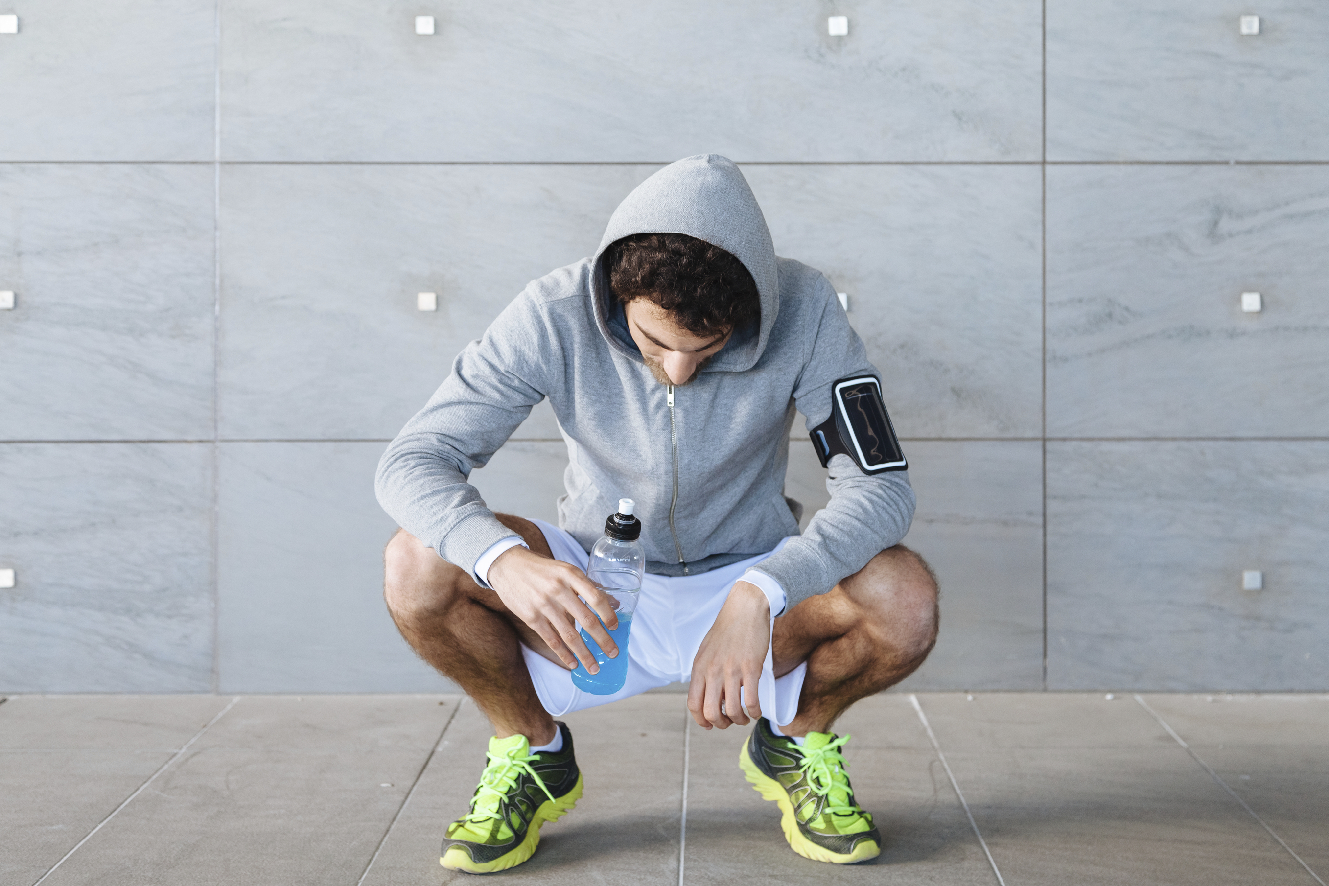 A young man in sportswear resting in the shade of a building in a summer day, drinking an energy drink blue color from a bottle before resuming training